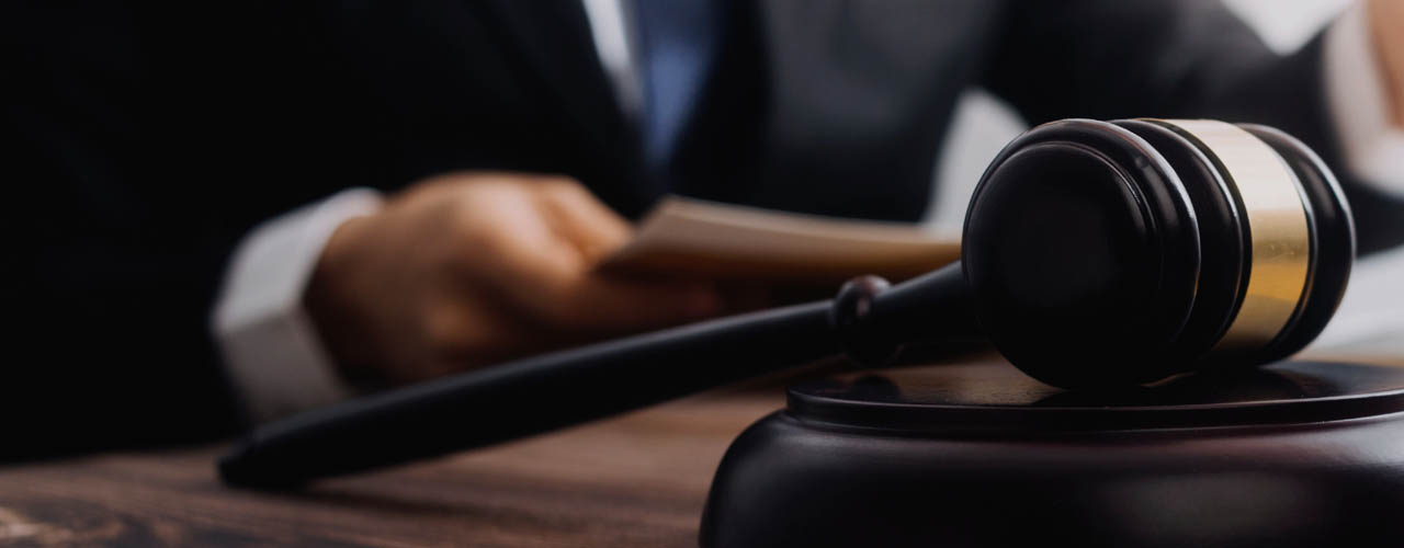 A gavel resting on a sounding block. A man in a suit is sitting at a desk in the background.