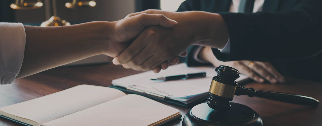 Two people shake hands over a desk. Papers, a gavel, and a set of scales sit on the desk.