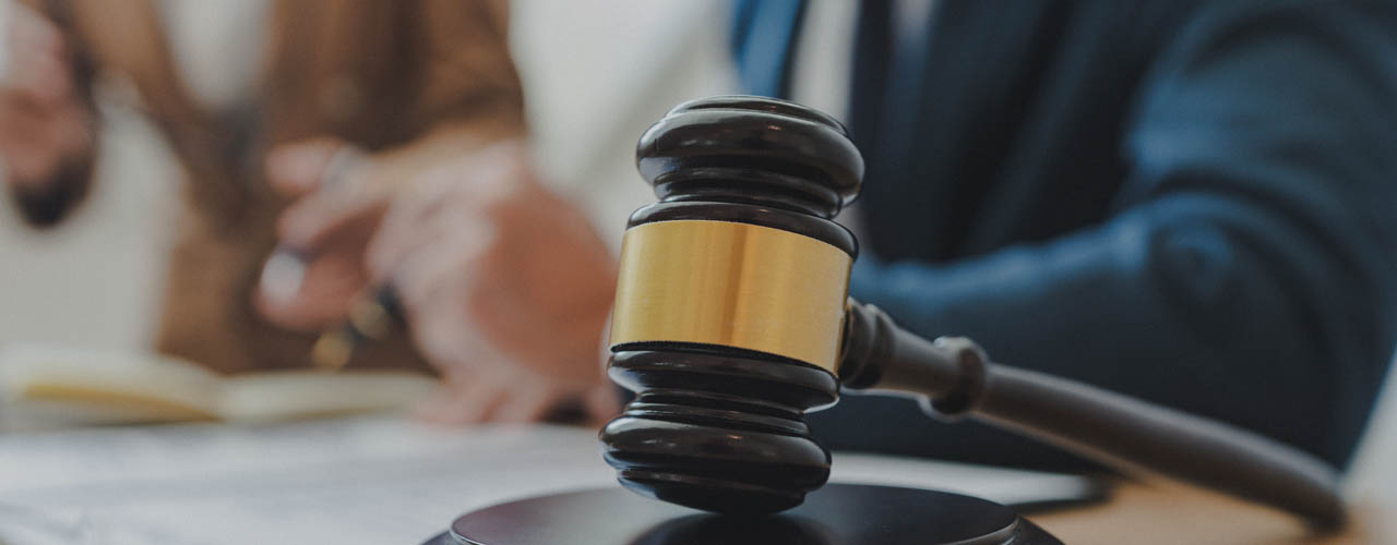 a gavel resting against a sounding block with two people sitting at a desk in the background.